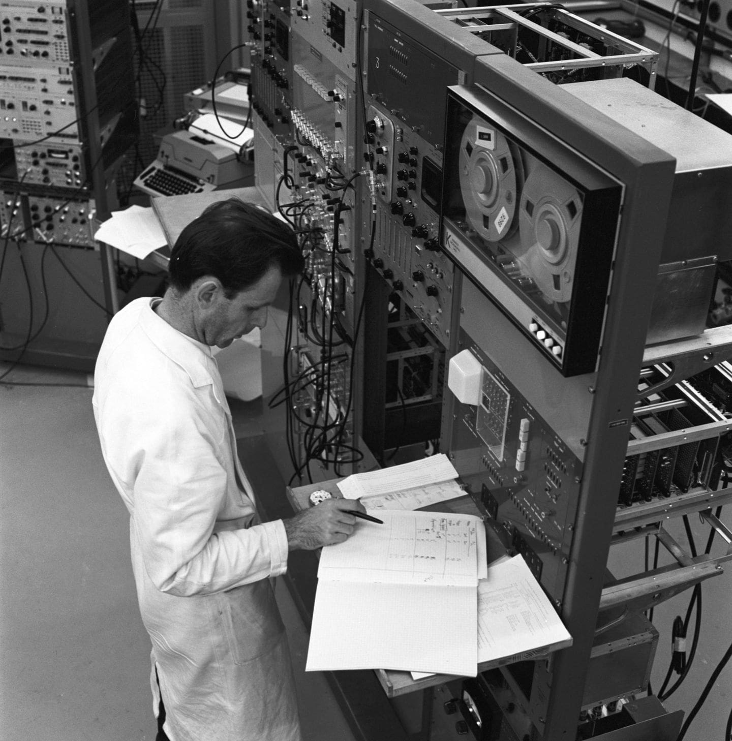 Man in white lab coat writing in a log book surrounded by machines
