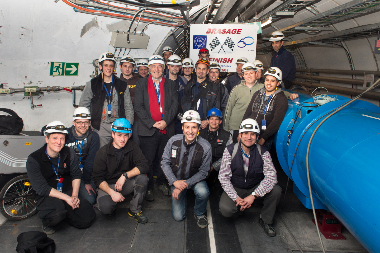 CERN engineers and technicians in hard hats pose for a photo in the LHC tunnel. The blue tube of the accelerator is on the right