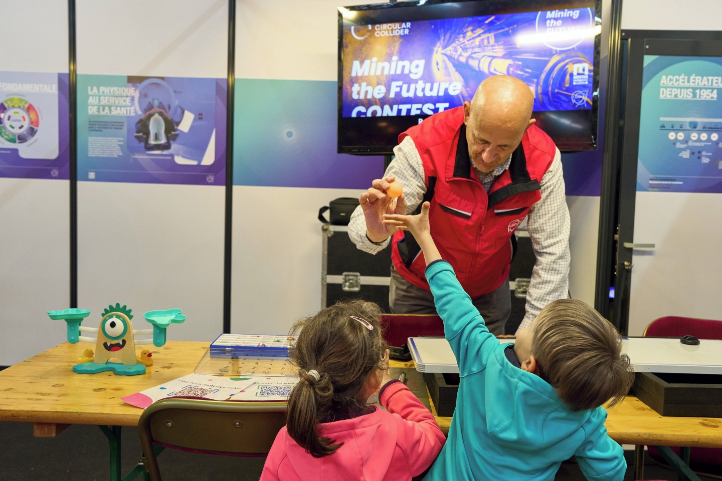 2 children in front of a table behind which a man is holding a ping-pong ball