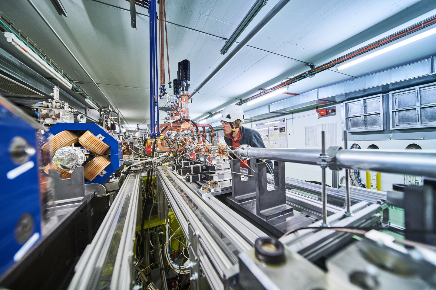 Young physicist with a helmet looking at a beam line equipped with magnets and copper structures