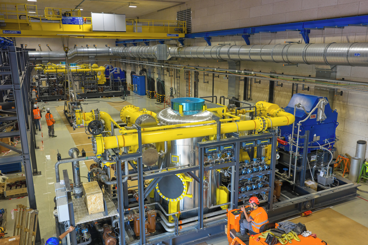 A complex structure with a silver cylinder, yellow big pipes and valves, is being moved by operators in a big industrial hall