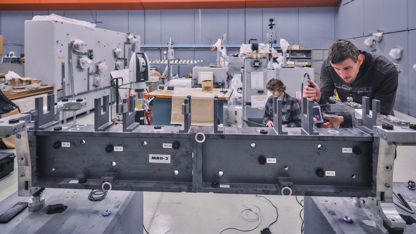 In the foreground is a large grey beam over which a man is leaning and looking at closely while holding some sort of sensor. In the background another man is sat with a laptop taking the data.